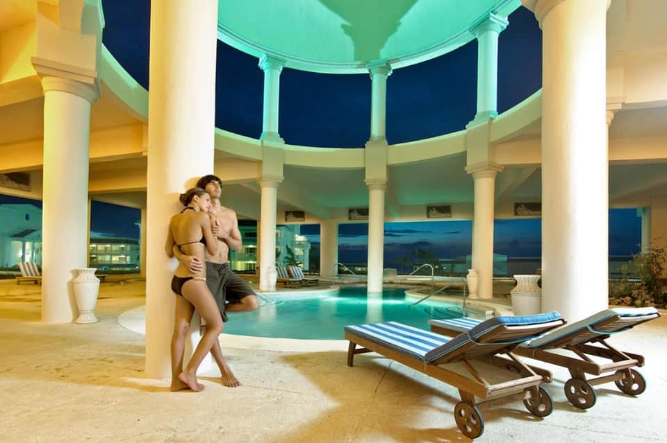 Couple by illuminated indoor pool with columns and night city views through curved windows