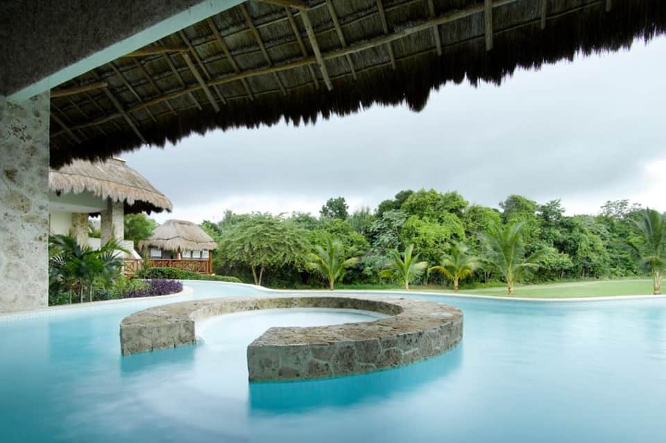 Luxury resort infinity pool with curved stone features, thatched-roof pavilion, and lush tropical landscape