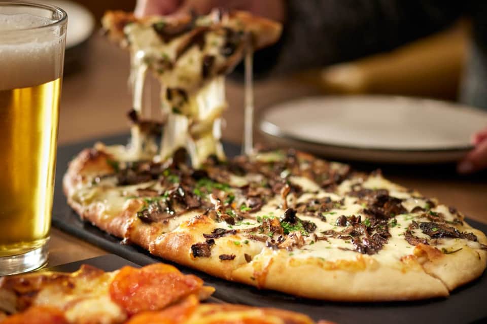 Flatbread pizza with meat and herbs served with beer on dark table at casual dining establishment