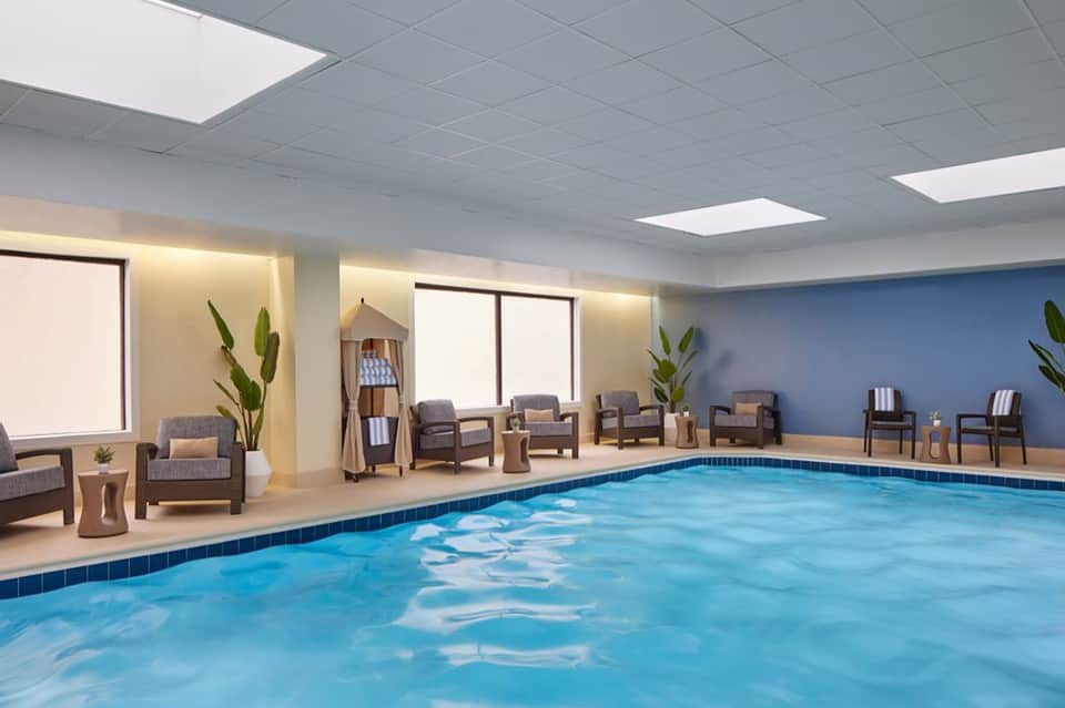 Indoor hotel pool with blue water, comfortable seating area, potted plants, and large windows with natural lighting