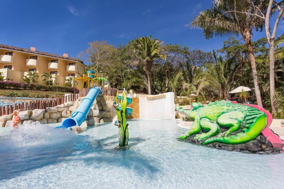 Colorful children's water park with blue slide, green crocodile sculpture, and tropical palm trees
