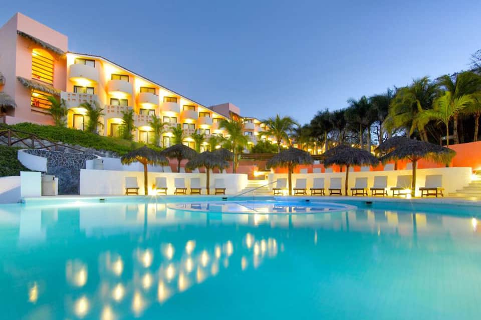 Illuminated resort building at dusk overlooking a turquoise pool with palm trees and lounge chairs
