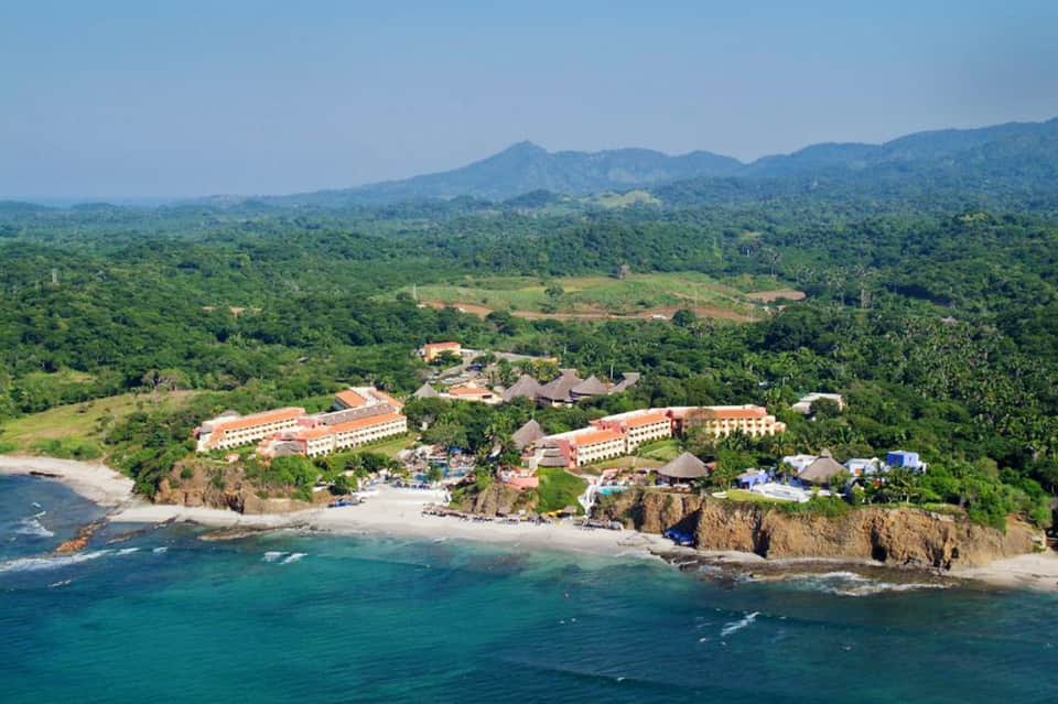 Aerial view of beachfront resort with orange-roofed buildings nestled between lush green mountains and turquoise ocean