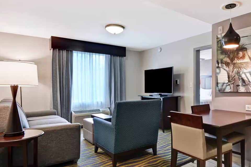 Modern hotel suite with seating area, TV, work desk, and floor-to-ceiling windows with gray curtains