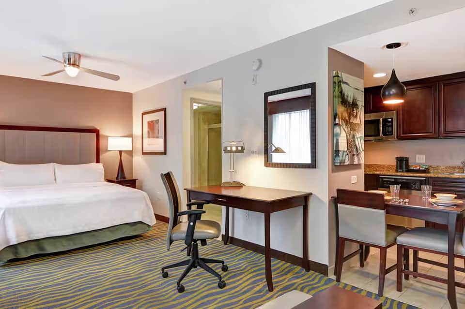 Modern hotel suite with queen bed, work desk, kitchenette, and striped area rug in warm tones