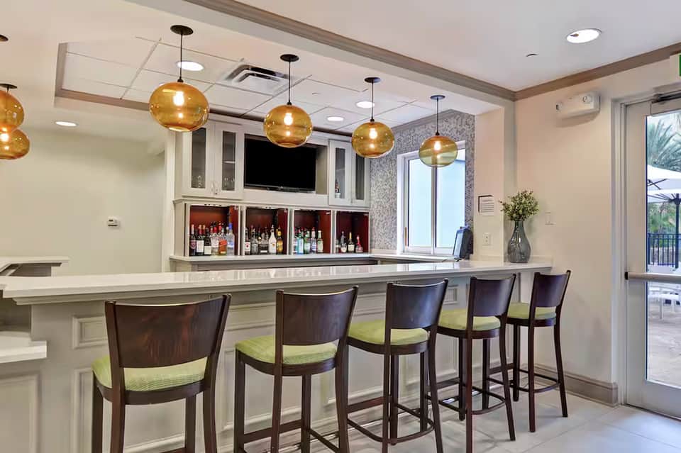 Modern resort bar with gold pendant lights, white marble countertop, dark wooden barstools with green cushions, and illuminated liquor display