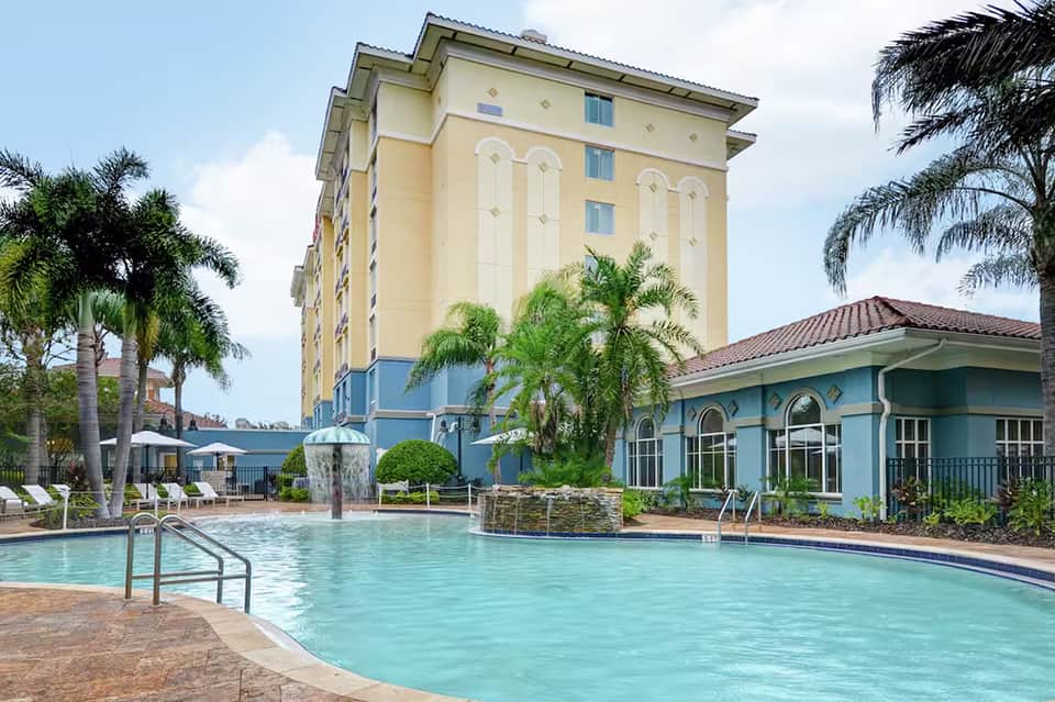 Yellow hotel building with pool, cabanas, and blue poolside structure surrounded by palm trees