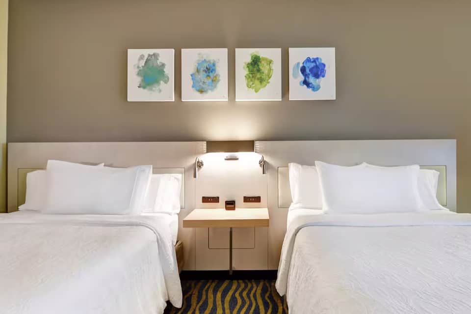 Modern hotel room with two queen beds, white linens, neutral headboard, and four colorful abstract watercolor paintings above