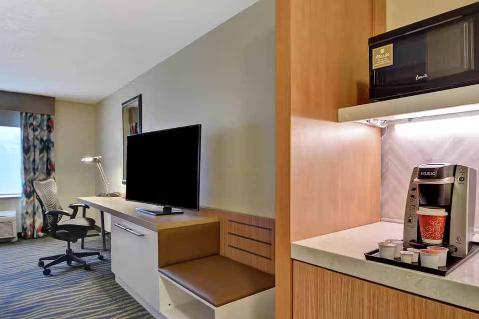 Modern hotel room with work desk, flat-screen TV, coffee maker, and wood cabinetry in neutral tones