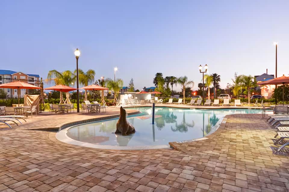 Resort pool deck at dusk with lounge chairs, umbrellas, palm trees, and lit lamp posts reflecting in calm water