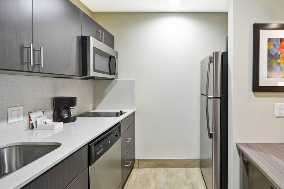 Contemporary hotel kitchenette with stainless steel appliances, dark cabinetry, and white countertops
