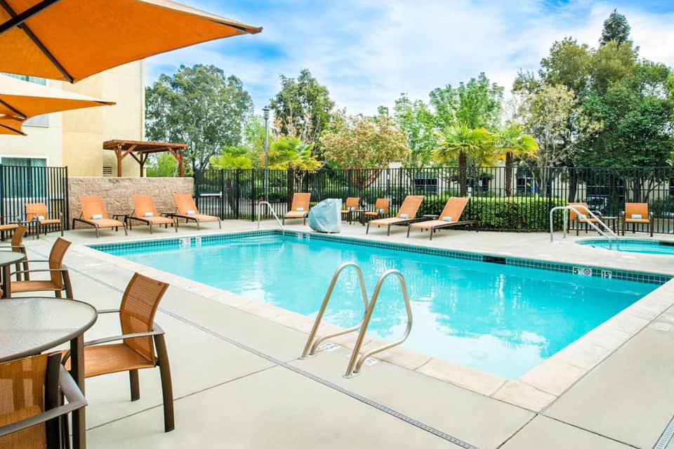 Resort pool with lounge chairs under orange umbrellas, surrounded by lush palm trees and manicured landscaping
