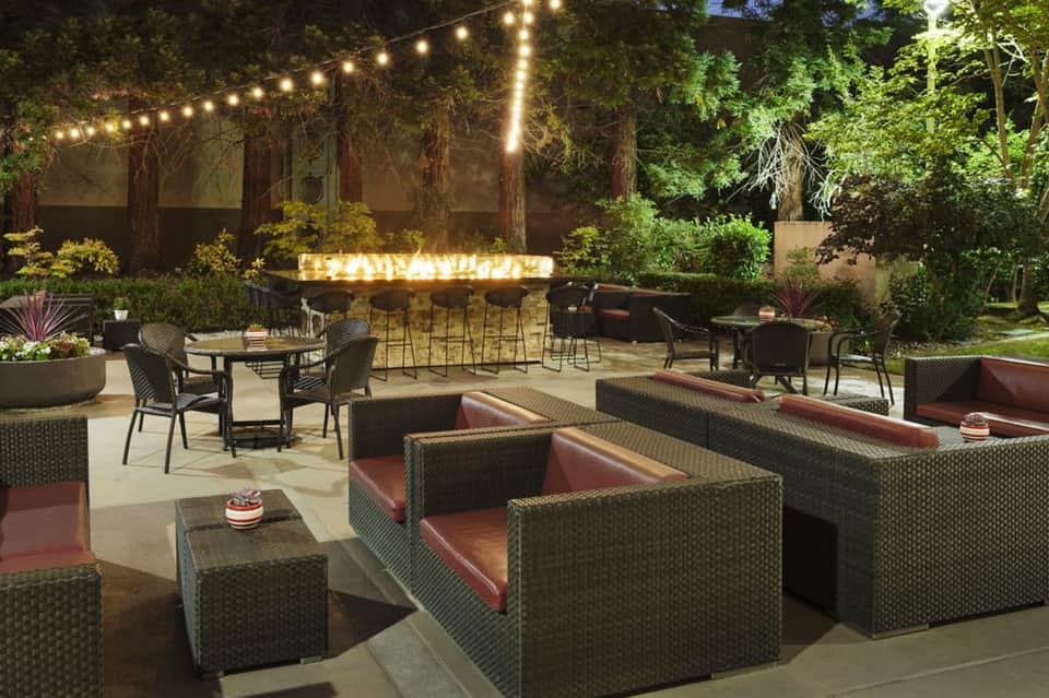 Outdoor patio at night with string lights, woven lounge seating with burgundy cushions, and illuminated bar in background