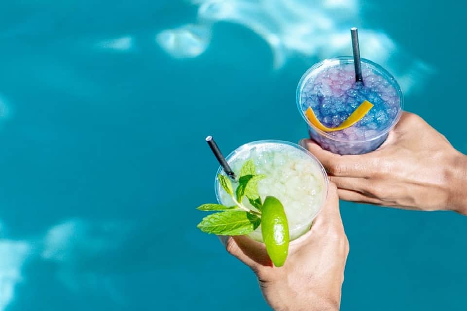 Two colorful tropical cocktails with straws held over turquoise water background