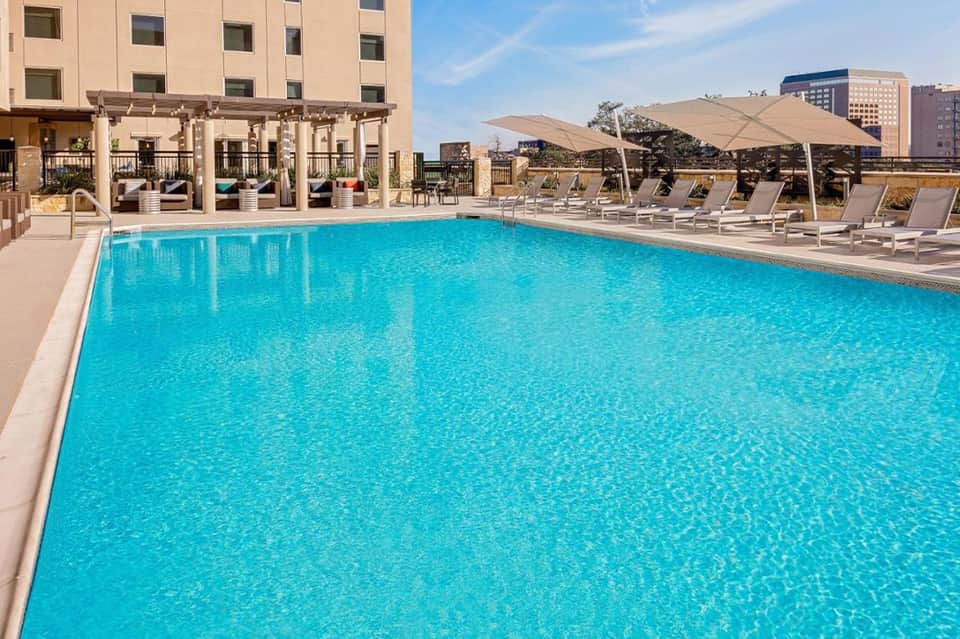 Large resort pool with crystal blue water, lounge chairs, umbrellas, and beige pergola structure with city buildings in background