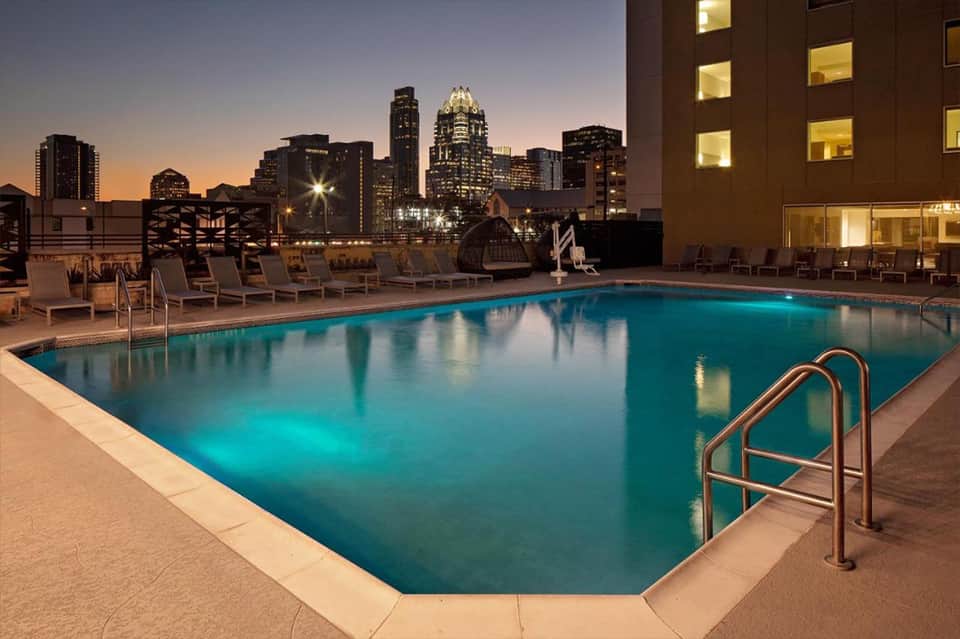 Illuminated rooftop pool at dusk with city skyline and lounge chairs