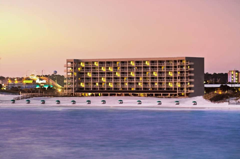 Beachfront hotel building at dusk with illuminated windows and beach umbrellas along the shore