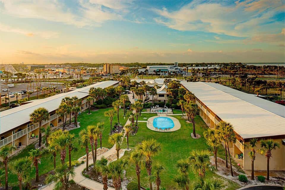 Aerial view of resort with central pool, palm trees, surrounding buildings, and coastal landscape at sunset