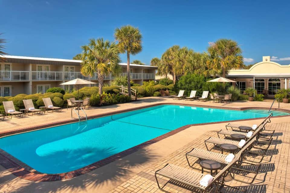 Resort pool with turquoise water, lounge chairs, palm trees, and two-story buildings under clear blue sky