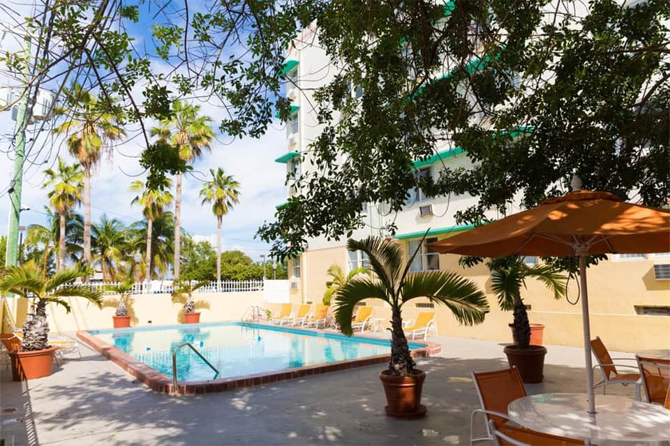 Resort pool surrounded by palm trees with yellow lounge chairs and tan umbrellas under clear sky