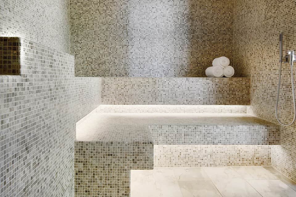 Luxury bathroom with mosaic tile walls, sunken bathtub, and rolled towels on shelf