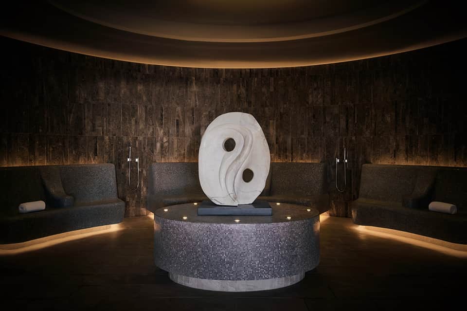 Luxurious spa interior with white yin-yang sculpture, dark stone walls, and ambient lighting, flanked by spa lounges