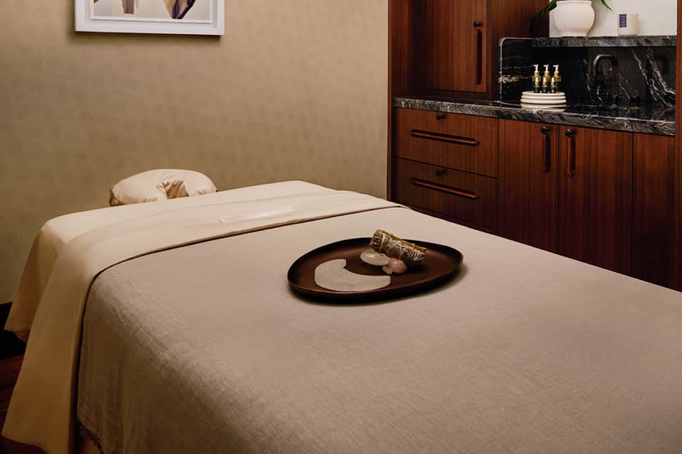 Spa treatment room with massage table, cream linens, dark wood cabinetry, and black marble countertop with decorative accessories