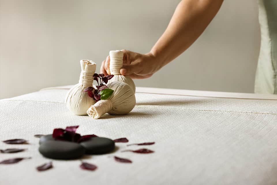 Hands holding a cream-colored herbal compress ball with dried flowers over a spa table