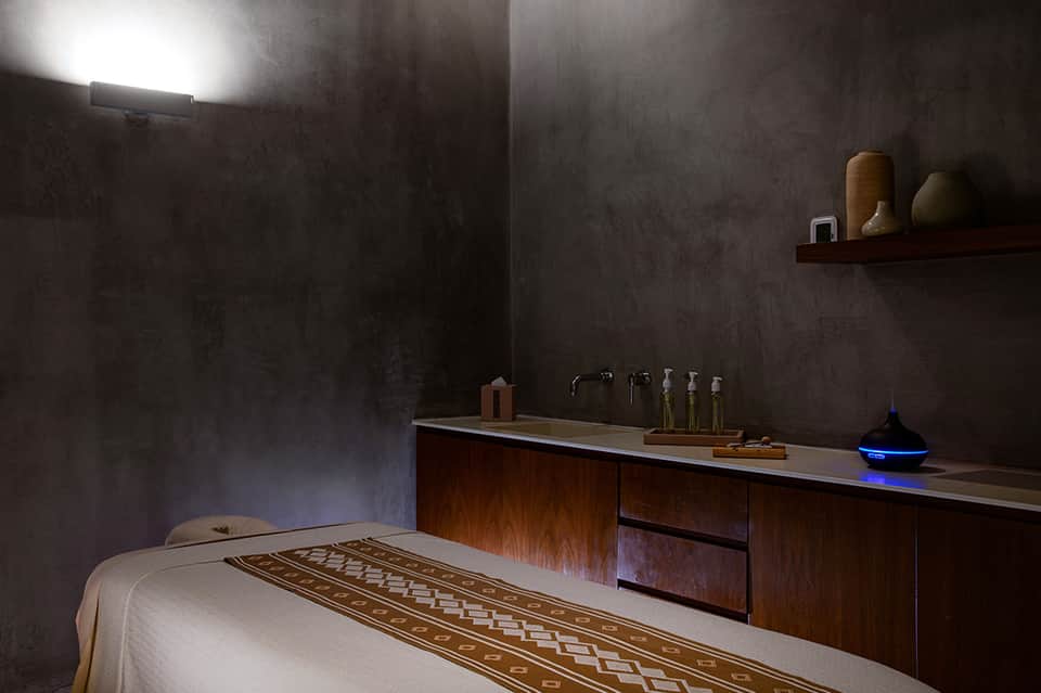Spa treatment room with massage table, dark textured walls, wooden vanity with oils and toiletries, and ambient lighting