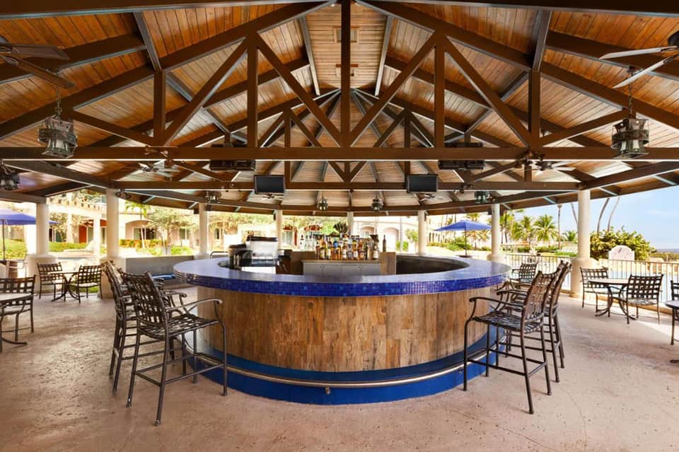 Large circular outdoor bar with blue tile rim under wooden truss ceiling, surrounded by lounge chairs