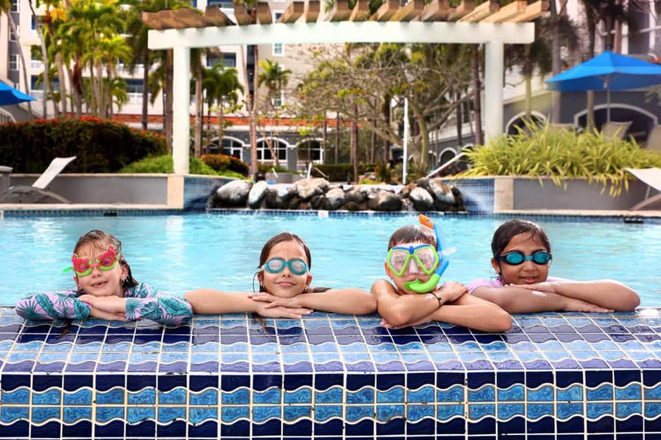 Four children wearing colorful snorkels at pool edge with resort buildings and umbrellas in background