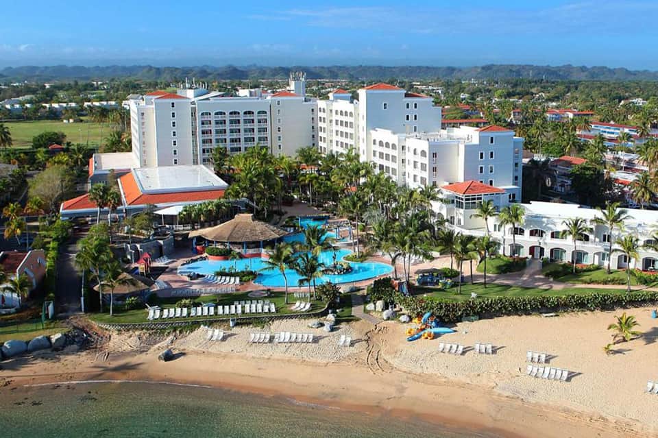 Beachfront resort with white multi-story buildings, blue pool complex, and sandy beach with lounge chairs