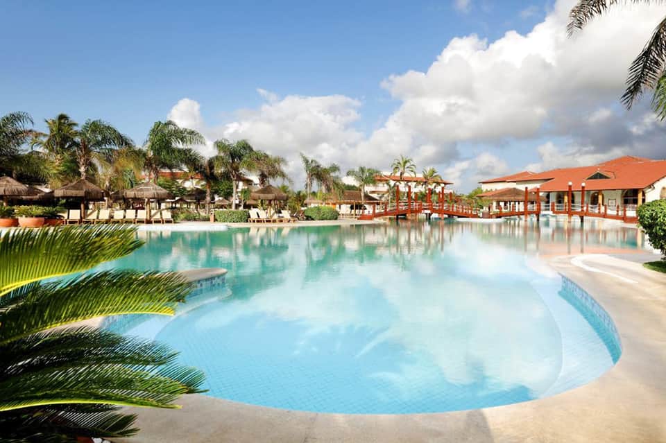 Expansive tropical resort pool with turquoise water, palm trees, orange-roofed buildings, and manicured grounds