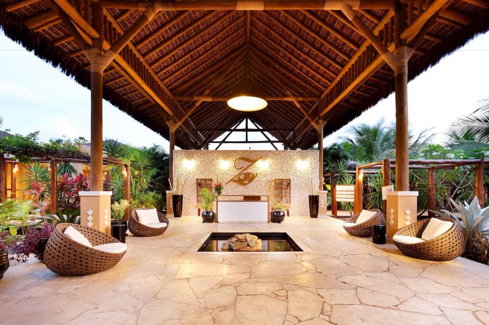 Luxury resort spa pavilion with wooden architecture, woven chairs, and stone fireplace surrounded by tropical plants