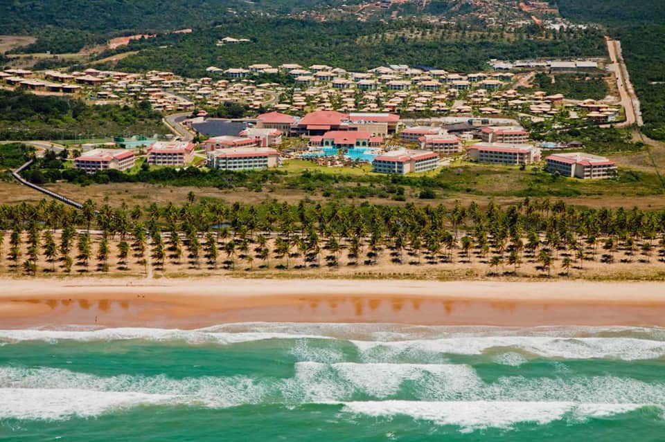 Aerial view of beachfront resort with red-roofed buildings, palm tree plantation, and ocean waves