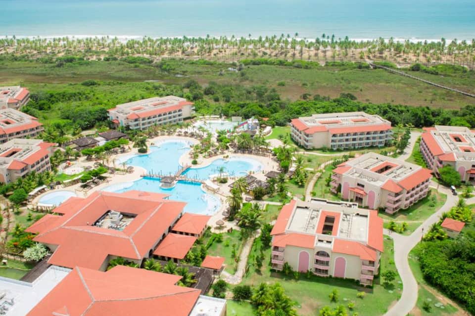 Aerial view of resort with red-roofed buildings, multiple pools, and palm trees near coastline