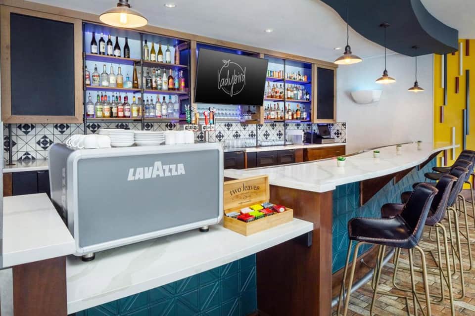 Contemporary hotel bar and lounge area with geometric tile backsplash, bottle display shelves, and blue accent seating