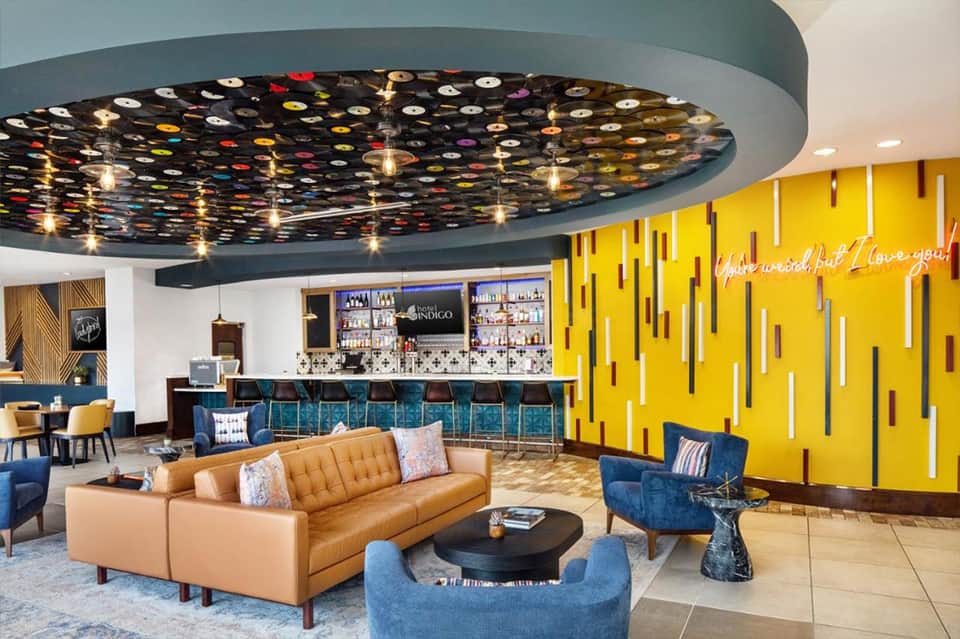 Modern hotel lobby with colorful ceiling installation, tan leather sofa, blue armchairs, yellow accent wall, and bar area