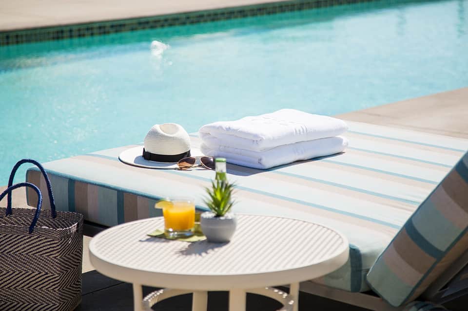 Poolside table with white towels, hat, sunglasses, and tropical drink on striped lounger deck