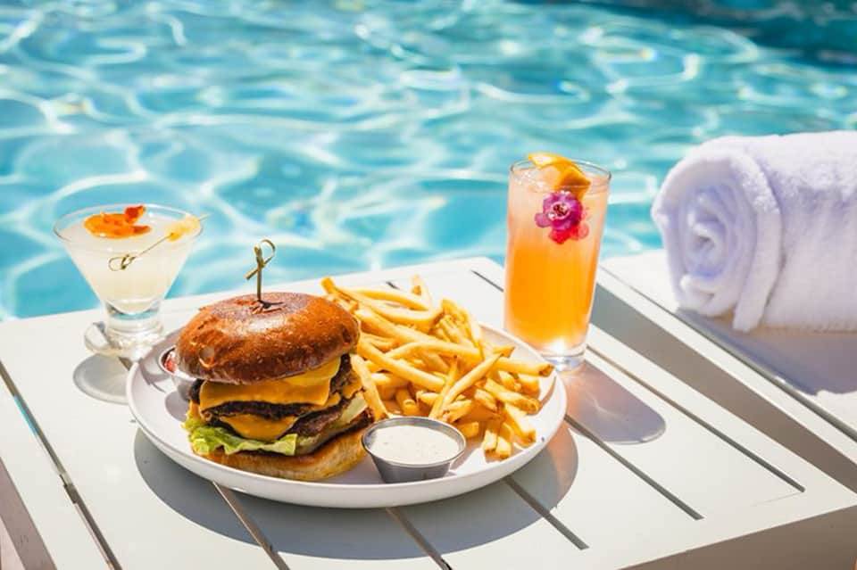 Gourmet burger with fries and cocktails poolside on white deck next to towel