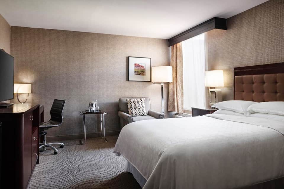 Modern hotel bedroom with brown upholstered headboard, white bedding, desk area, and neutral textured wallpaper