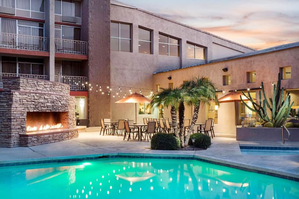 Modern resort pool at dusk with fireplace, dining area, palm trees, and contemporary architecture