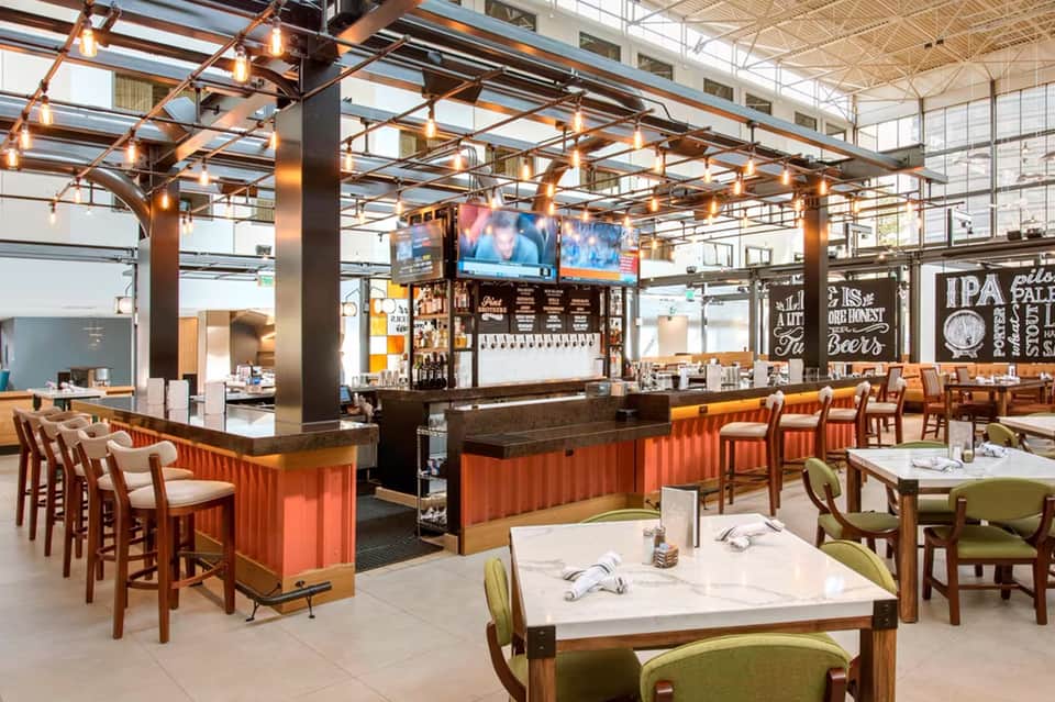 Modern brewery restaurant with orange corrugated bar, wooden chairs, high ceilings with copper pendant lights, and dining tables