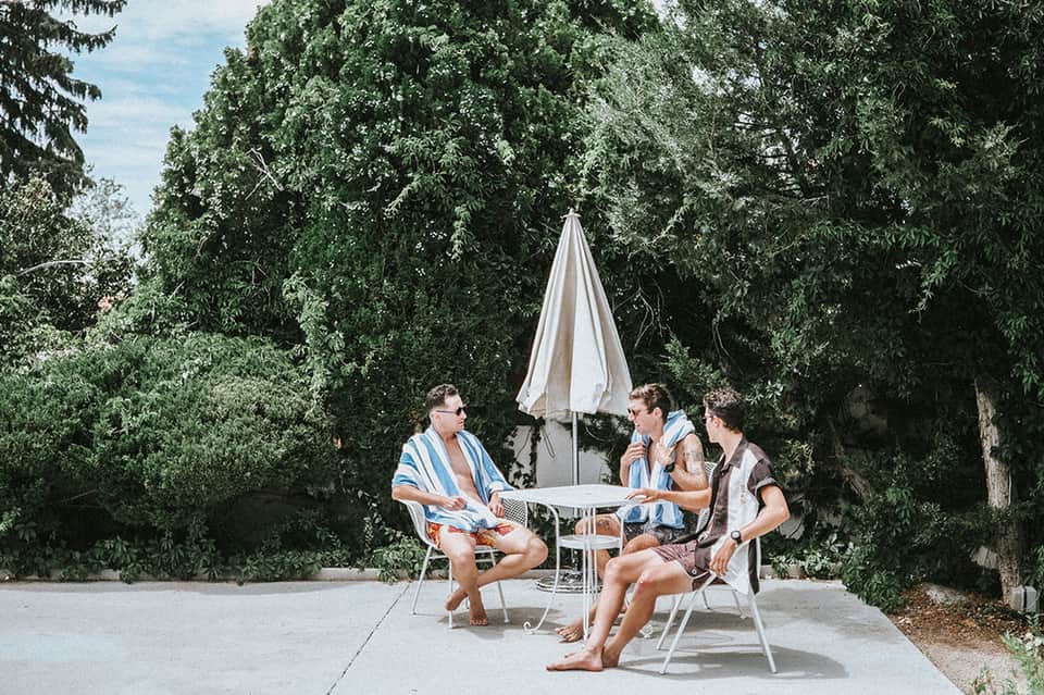 Three men relaxing under umbrella in resort courtyard surrounded by tall green trees