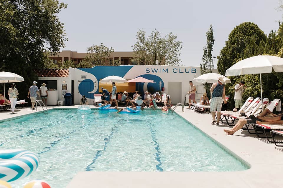Busy swim club with rectangular pool, white umbrellas, lounge chairs, and blue clubhouse building
