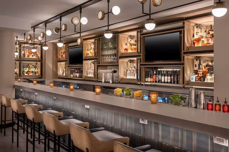 Contemporary bar interior with brass pendant lights, wooden shelving displaying liquor bottles, TV screens, and tan bar stools