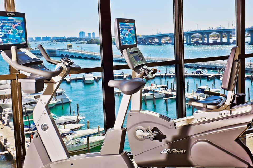 Exercise bikes by large windows overlooking blue harbor, bridge, and marina with boats