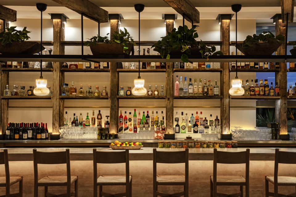 Upscale bar with wooden beams, stocked shelves of spirits, pendant lighting, and potted plants along the counter