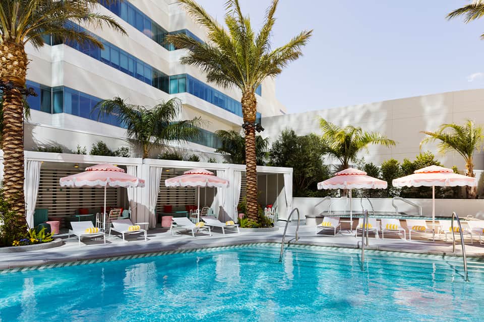Resort pool with crystal-clear turquoise water, pink umbrellas, lounge chairs, and white cabanas with modern white building.