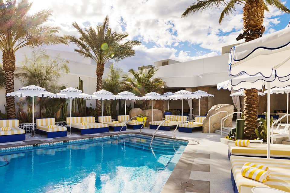 Resort pool deck with blue water, palm trees, yellow and white striped loungers, white umbrellas, and clear sky.